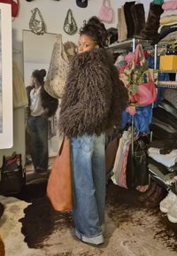 Barna, bolyhos, túlméretezett kabát, világos farmer, barna kézitáska; a tükör visszatükrözi egy nő képét egy zsúfolt szobában, különféle táskák és ruhák között.