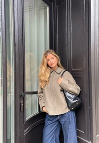 Világosbarna, cipzáros dzseki széles ujjal, sötétkék, széles lábú farmerral párosítva. Fekete bőr válltáska kontrasztot ad. Geometrikus ajtókeret háttér.