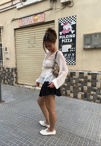 Valkoinen, läpikuultava pitkähihaisuus pusero mustan pilkullisen mini-hameen päällä. Asusteena valkoiset ballerinat. Taustalla on pizzerian kuva.