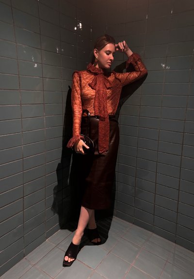 Blusa de encaje color óxido con cuello anudado, combinada con una falda de cuero negra. Zapatos de tacón abiertos negros, sosteniendo un bolso pequeño. Azulejos texturizados de fondo.
