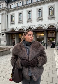 Grå konstpälsjacka, stickade bruna handskar, grå scarf och en mörkbrun handväska med dekorativa röda charmiga detaljer, placerade framför en vit byggnad.