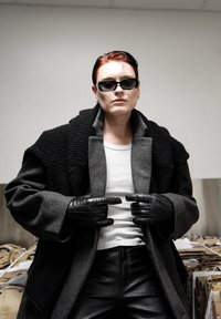 Guanti di pelle nera, cappotto grigio a trama con colletto spesso, camicia bianca e pantaloni in pelle nera. Sovrapposizioni sottili e elementi di design moderni.