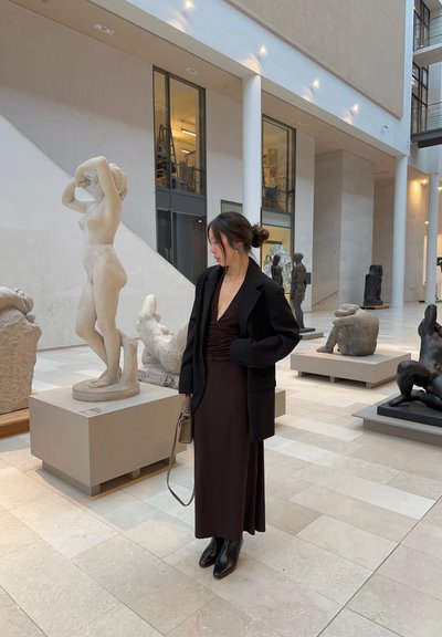 Blazer negro sobre un vestido ajustado marrón oscuro, combinado con botas de tobillo negras. En una galería con varias esculturas y un suelo de azulejos claros.