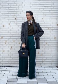 Kvinne med flettet hår som bærer et skjerf, rutete blazer, olivengrønn genser, vide Adidas-bukser, holder en svart veske, står ved en hvit murvegg.