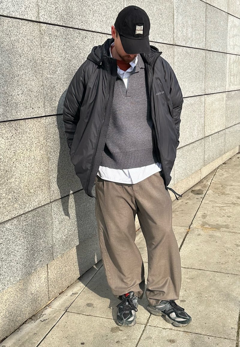 Homme portant une casquette noire, une veste noire, un pull gris, un pantalon beige et des baskets, debout contre un mur carrelé sur un trottoir ensoleillé.