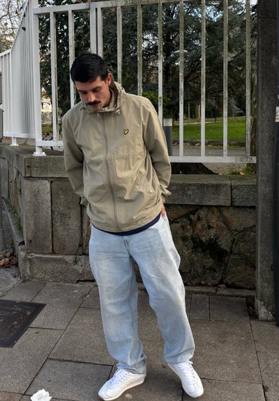 Chaqueta cremosa con cierre y un logo en el pecho, combinada con jeans de mezclilla azul claro y zapatillas blancas. De pie al aire libre cerca de una valla.