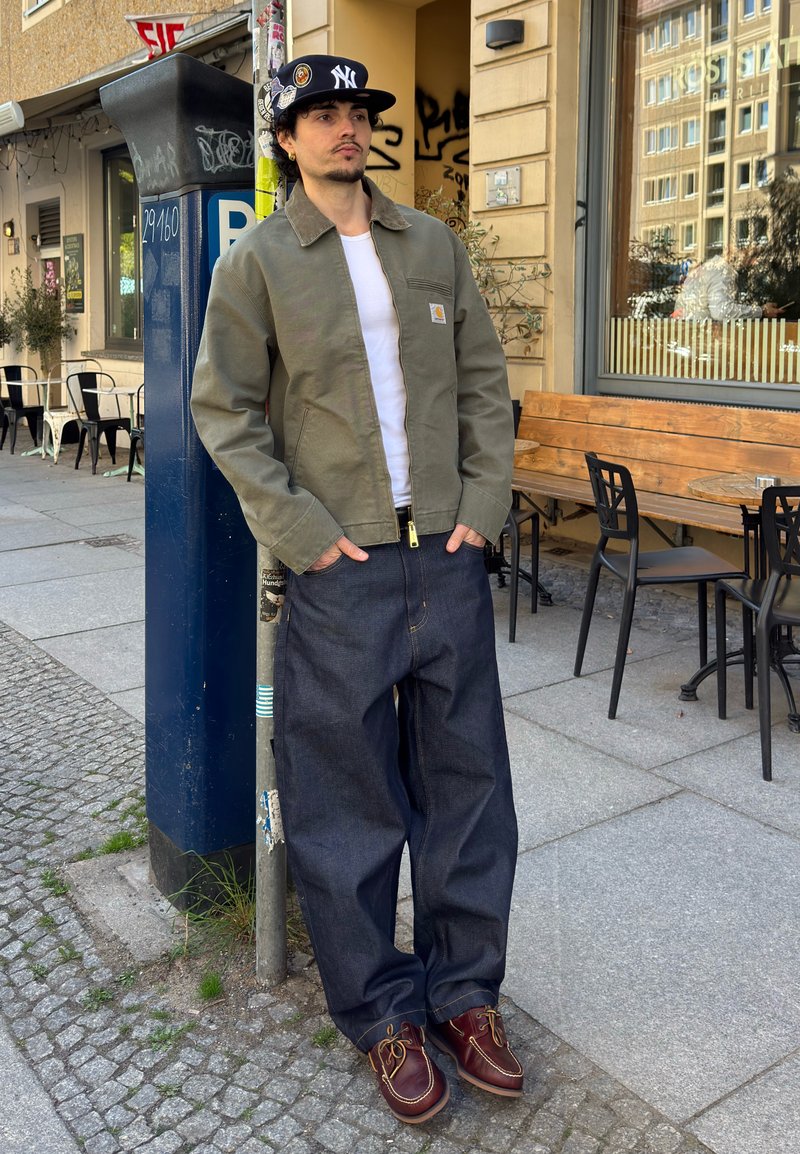 Jeune homme en tenue décontractée avec casquette appuyé contre un parcmètre près des tables d’un café sur un trottoir urbain.