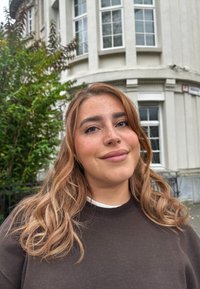 Ένα άτομο με μακριά, κυματιστά καστανά μαλλιά ντύνεται με ένα σκούρο πουλόβερ πάνω από ένα λευκό πουκάμισο και στέκεται έξω κοντά σε πράσινη βλάστηση και ένα κτίριο με μεγάλες τζαμαρίες.