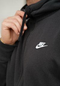 Hanorac cu fermoar negru, dintr-un material moale, prevăzut cu un logo Nike brodat alb pe piept și un sistem de reglare cu șnur la glugă.