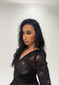 Zwart doorschijnend topje met ingewikkelde golfpatronen, lange mouwen en een diep decolleté. Draagt een zilveren hangersketting.