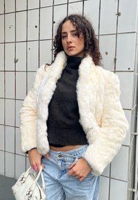 Valekunstnahast kooretooniline jope kõrge kraega, pikkade varrukatega ja tekstuurse kangaga, koos musta lühikese kampsuni ja heleroheliste teksadega.