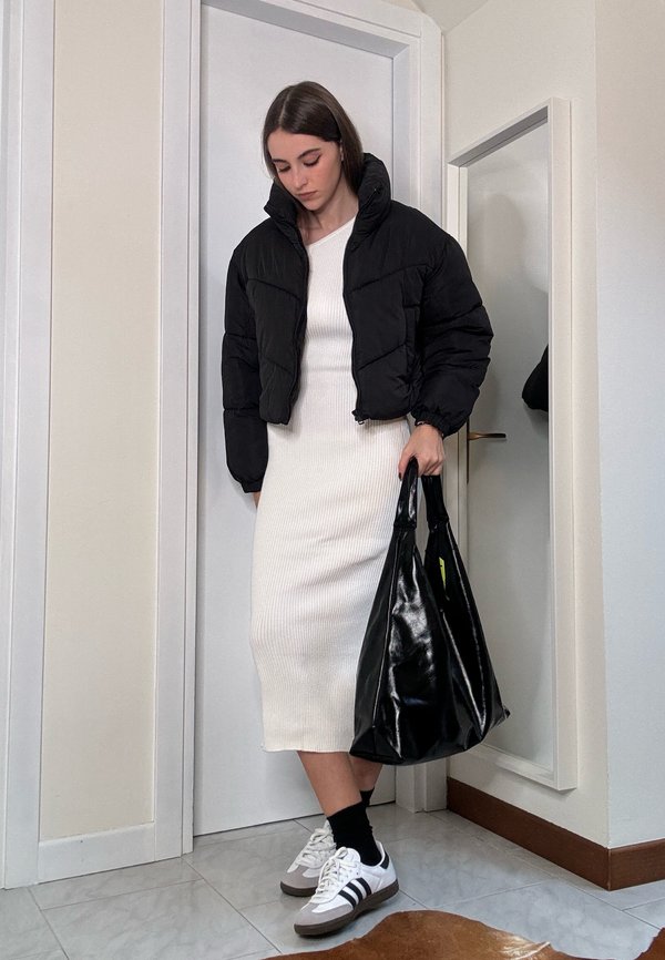 Casaco puffer preto, vestido midi branco com relevo, bolsa preta, sapatilhas cinza e brancas, usadas com meias pretas, de pé num ambiente interno simples.