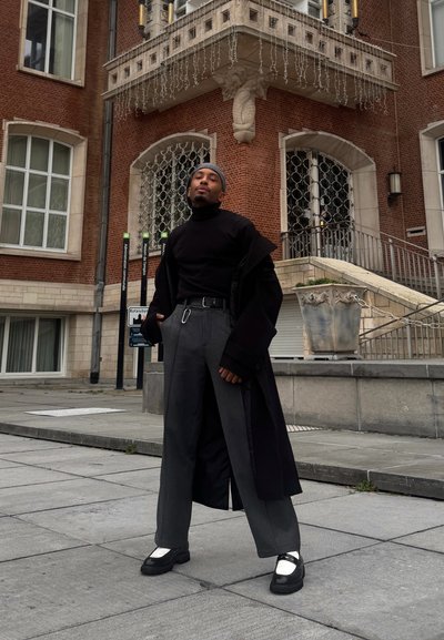 Cuello alto negro y pantalones anchos grises bajo un abrigo largo negro; zapatos negros con detalles metálicos; fondo urbano con edificio de ladrillo y escaleras.