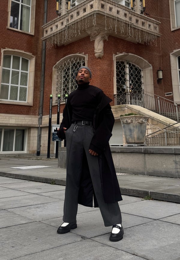 Svart polotröja och vida grå byxor under en lång svart kappa; svarta skor med metalliska detaljer; stadsbakgrund med tegelbyggnad och trappor.