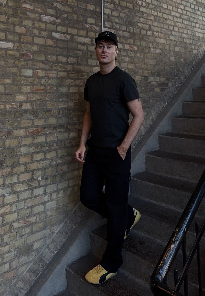 T-shirt noir à manches courtes, pantalon noir et baskets jaunes. Debout sur des escaliers, portant une casquette noire et avec des tatouages visibles sur le bras.