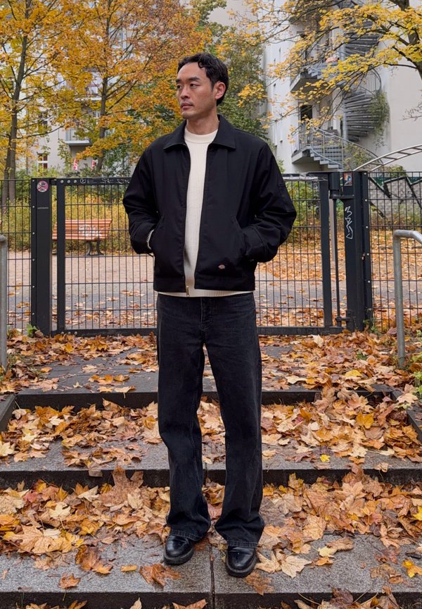 Un homme en veste noire et en jean foncé se tient, les mains dans les poches, parmi des feuilles mortes d’automne ; derrière lui se trouve une cour fermée par un portail.