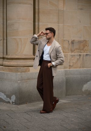 Blazer de color beige de doble pecho sobre una camisa blanca, pantalones anchos marrones, zapatos marrones, de pie frente a una pared de piedra con columnas.