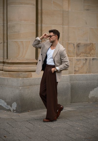 Blazer de color beige de doble pecho sobre una camisa blanca, pantalones anchos marrones, zapatos marrones, de pie frente a una pared de piedra con columnas.