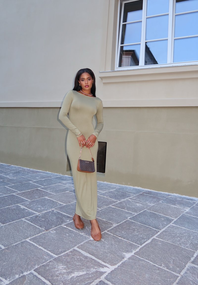 Πράσινο ελαιόλαδο μάξι φόρεμα, εφαρμοστό, με απαλή υφή; συνδυασμένο με καφέ τσάντα. Μοντέλο που φοράει καφέ φλατ, στέκεται σε πέτρινο δάπεδο.