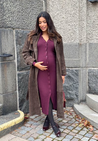 Un largo vestido burdeos de canalé con botones, llevado bajo un abrigo de ante marrón. Medias negras y zapatos de un burdeos oscuro completan el conjunto.