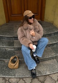 Műszőrme kabát zabkása színben, laza fazonú kék farmernadrág, fekete mokaszin arany díszítéssel, barna sapka felirattal, kávésbögrét tartva, szövött táska.