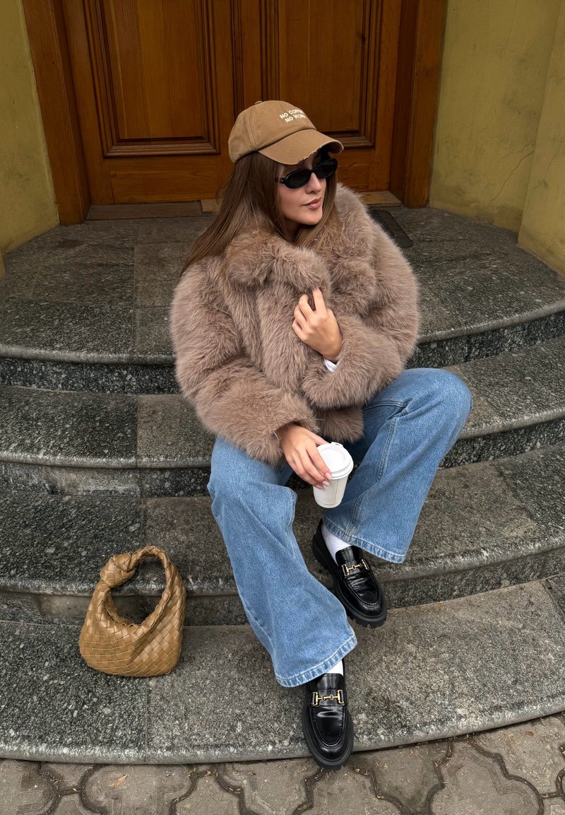 Manteau en fausse fourrure couleur avoine, jean bleu coupe décontractée, mocassins noirs avec ornements dorés, casquette marron avec texte, tenant une tasse de café, sac tissé.