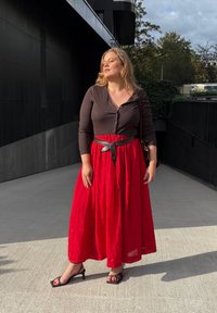 Žena s plavom kosom koja nosi crnu majicu, crvenu suknju, crni pojas i crne štikle stoji na otvorenom na suncu s zatvorenim očima.