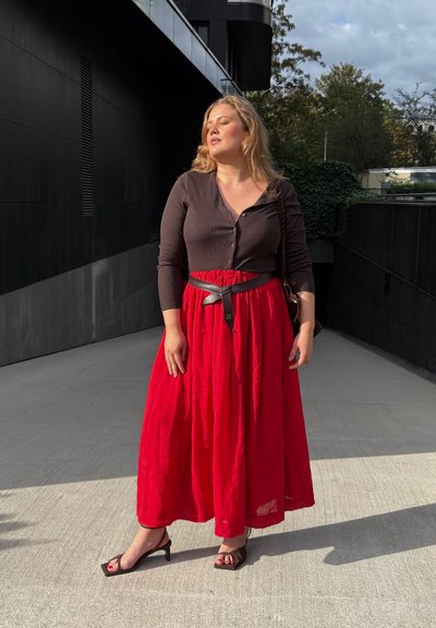 Mujer con cabello rubio que lleva una blusa negra, una falda roja, un cinturón negro y tacones negros, de pie al aire libre bajo la luz del sol con los ojos cerrados.