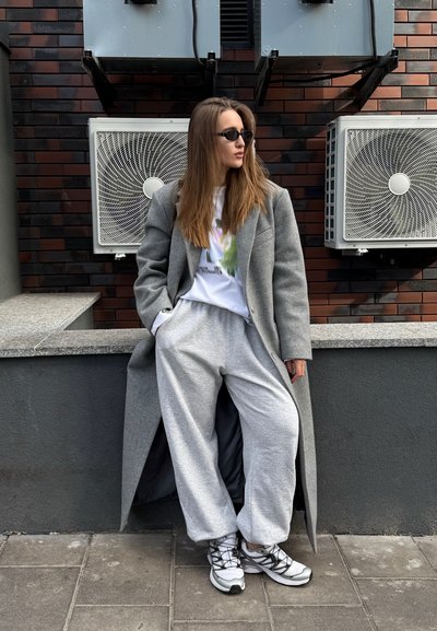 Abrigo de lana gris, camiseta gráfica blanca, pantalones de chándal gris holgados y zapatillas deportivas blancas. El entorno incluye unidades de aire acondicionado en una pared de ladrillos.