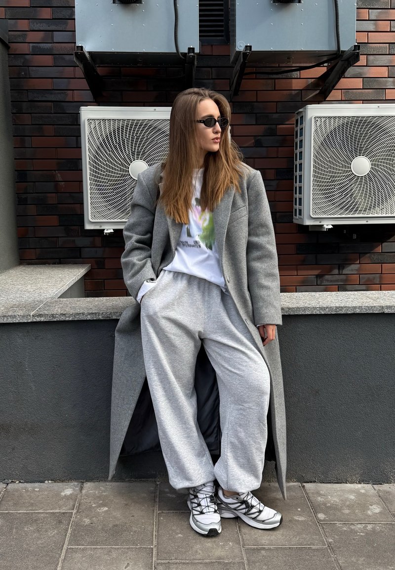 Gray wool coat, white graphic t-shirt, loose gray sweatpants, and white athletic shoes. The setting includes air conditioning units on a brick wall.