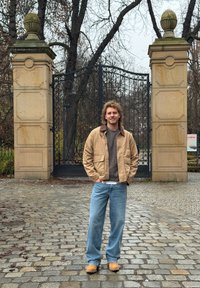 Veste en coton beige avec col marron sur pull gris, jean bleu à jambes droites et chaussures couleur beige, debout près d'une porte décorative.