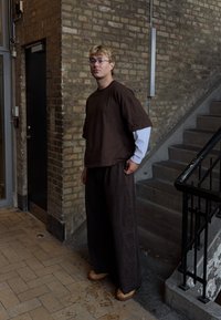 Haut ample marron et pantalon large assorti avec des accents texturés. Chemise blanche à manches longues en dessous. Sabots en bois aux pieds.