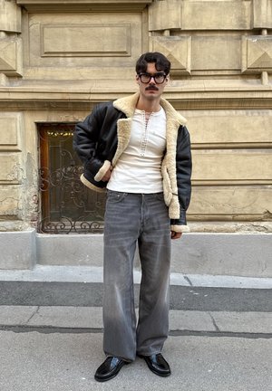 Chaqueta de shearling negra de cuero, camisa blanca ajustada con detalle de botones, jeans gris de pierna ancha y zapatos negros brillantes. Fondo urbano.