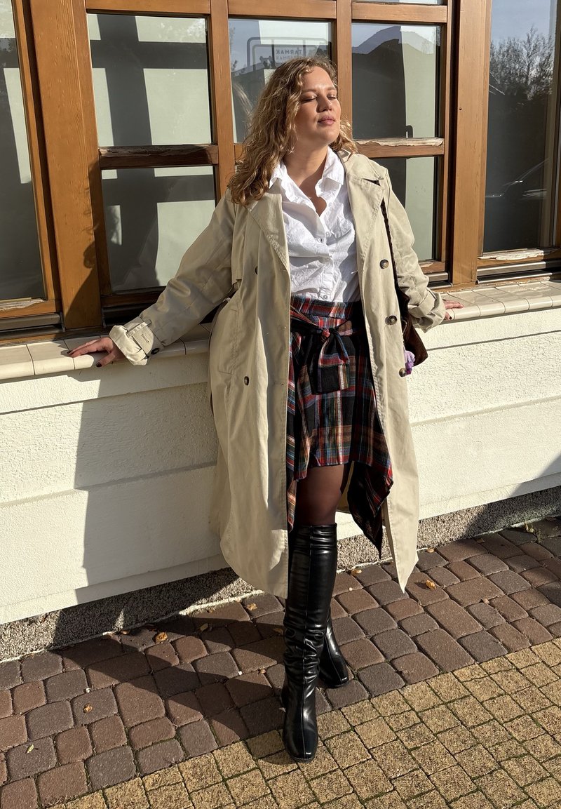 Beige trenchcoat over een wit shirt, gecombineerd met een geruite rok, zwarte kniehoge laarzen en lang, krullend haar. Zonschijn werpt zachte schaduwen.