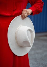 Biely plstený kovbojský klobúk s prehnutou korunou a dekoračným pásom. Držaný rukou s obväzom, pred červeným oblečením.