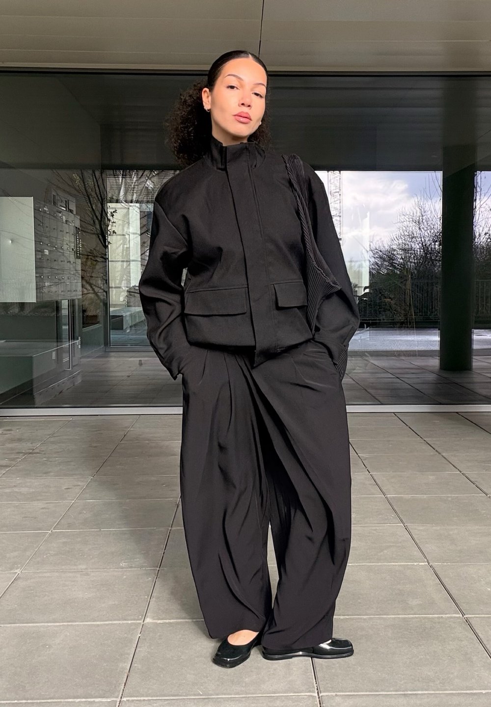 Sort jakke med høj krave og to forlommer, kombineret med vide sorte bukser. Glat stof med minimal detaljering. Sorte flade sko.