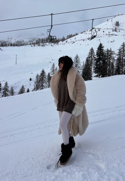 Abrigo de piel suave sobre un sudadera marrón, leggins blancos y botas negras. Paisaje cubierto de nieve con un telesilla al fondo.