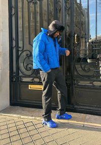 Blå dunjakke med hætte, sorte jeans og blå sneakers. Jakken har en quiltet tekstur; baggrunden viser en udsmykket sort port.