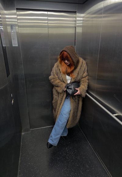 Abrigo de piel marrón con capucha, vaqueros de talle alto azul claro y botas negras con tacón. Bolso negro. Fondo de ascensor metálico.