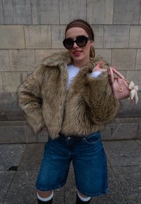 Τεχνητό γουναρικό κοντό μπουφάν σε καφέ απόχρωση, τζιν σορτσάκια και μαύρες μπότες μέχρι το γόνατο. Ρόζ τσάντα χειρός με λεπτομέρεια φιόγκο. Λευκό πουκάμισο με μακριά μανίκια από κάτω.