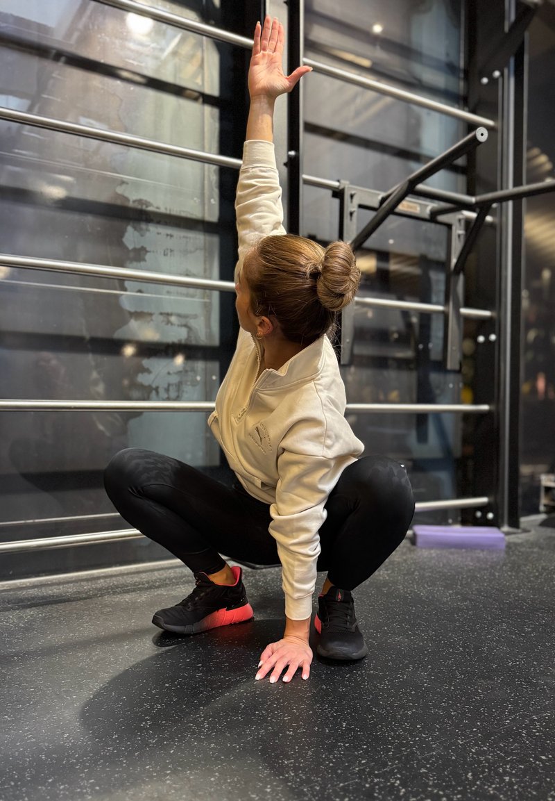 Athlète portant un sweat à capuche à fermeture éclair de couleur claire et des leggings noirs, accroupie sur le sol de la salle de sport, avec un bras étendu vers le haut. Chaussures de sport noires avec des accents rouges.