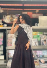 Kvinde med krøllet hår og briller, der retter på sit hår, iført en sort kjole, grå tørklæde og en sort brysttaske i en butiksindstilling.