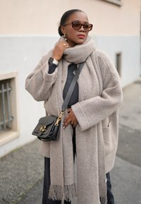 Μπεζ κασμίρ κάρντιγκαν με υπερμεγέθη μανίκια, σε συνδυασμό με γκρι μαντήλι με φούντες. Μαύρη τσάντα χεριού με χρυσές λεπτομέρειες και υφή.