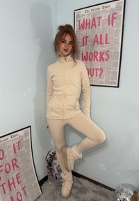 Bež flis jakna in ujemajoče oprijete hlače s visokim ovratnikom, žepki z zadrgo in obutvijo z mehkim poudarkom. V ozadju je okvirjeno besedilo.