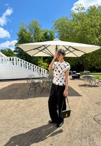 Biała bluzka w czarne kropki, zaprojektowana w stylu luźnym; luźne czarne spodnie; czarna torebka; plener z dużym parasolem i krzesłami z kutego żelaza.