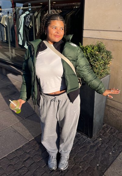 Chaqueta de plumas verde sobre un suéter negro, top corto blanco y pantalones de chándal grises. Sosteniendo una bebida, se encuentra frente a la ventana de una tienda.