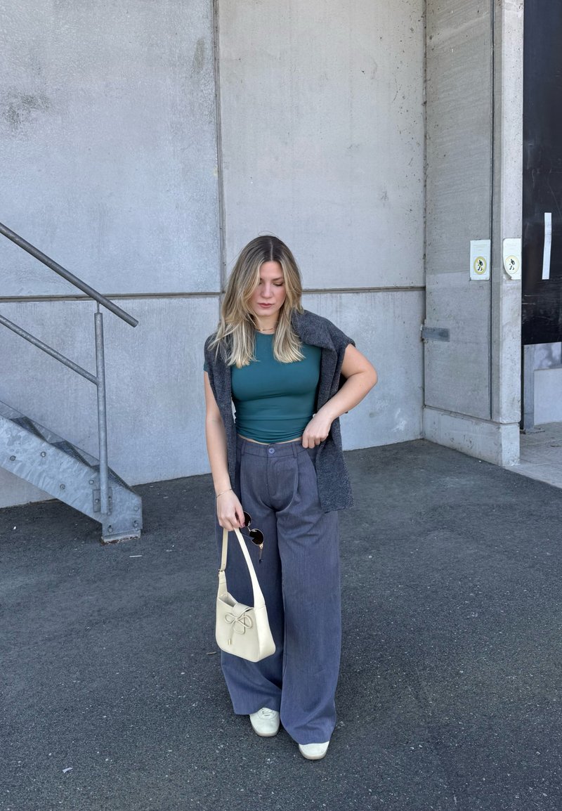 Femme aux longs cheveux blonds portant un haut turquoise, un gilet gris, un pantalon large gris, des chaussures blanches, tenant un sac à main crème et des lunettes de soleil près d'un mur en béton et des escaliers.