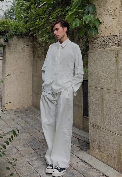 Camisa blanca de rayas verticales, de corte holgado y con mangas largas, combinada con pantalones blancos sueltos y zapatillas negras, todo ello con un fondo verde.