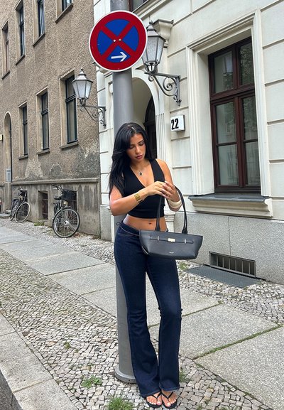 Bolso negro con un diseño elegante, llevado con jeans acampanados oscuros y una blusa ajustada negra, de pie en una calle de piedra cerca de un cartel.