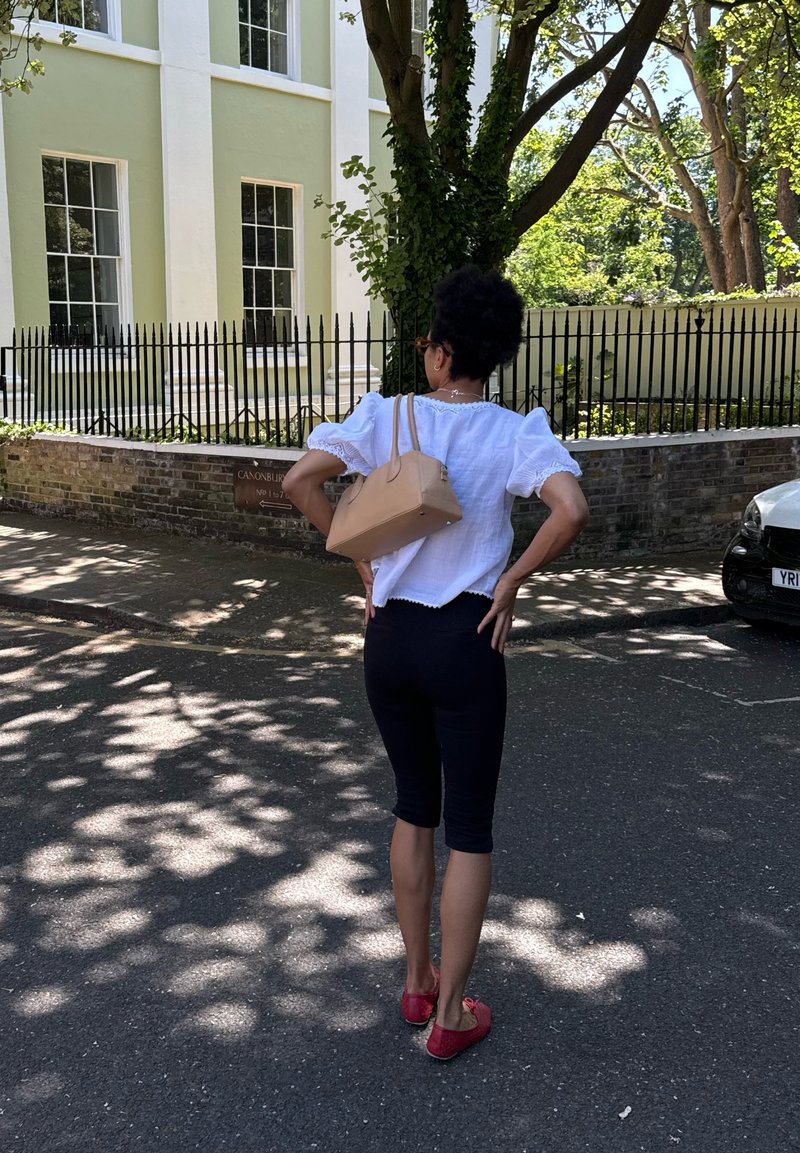 Femme aux cheveux courts et bouclés, vêtue d'un chemisier blanc et d'un pantalon capri noir, debout les mains sur les hanches dans une rue ensoleillée près d'un bâtiment jaune et d'une clôture noire.